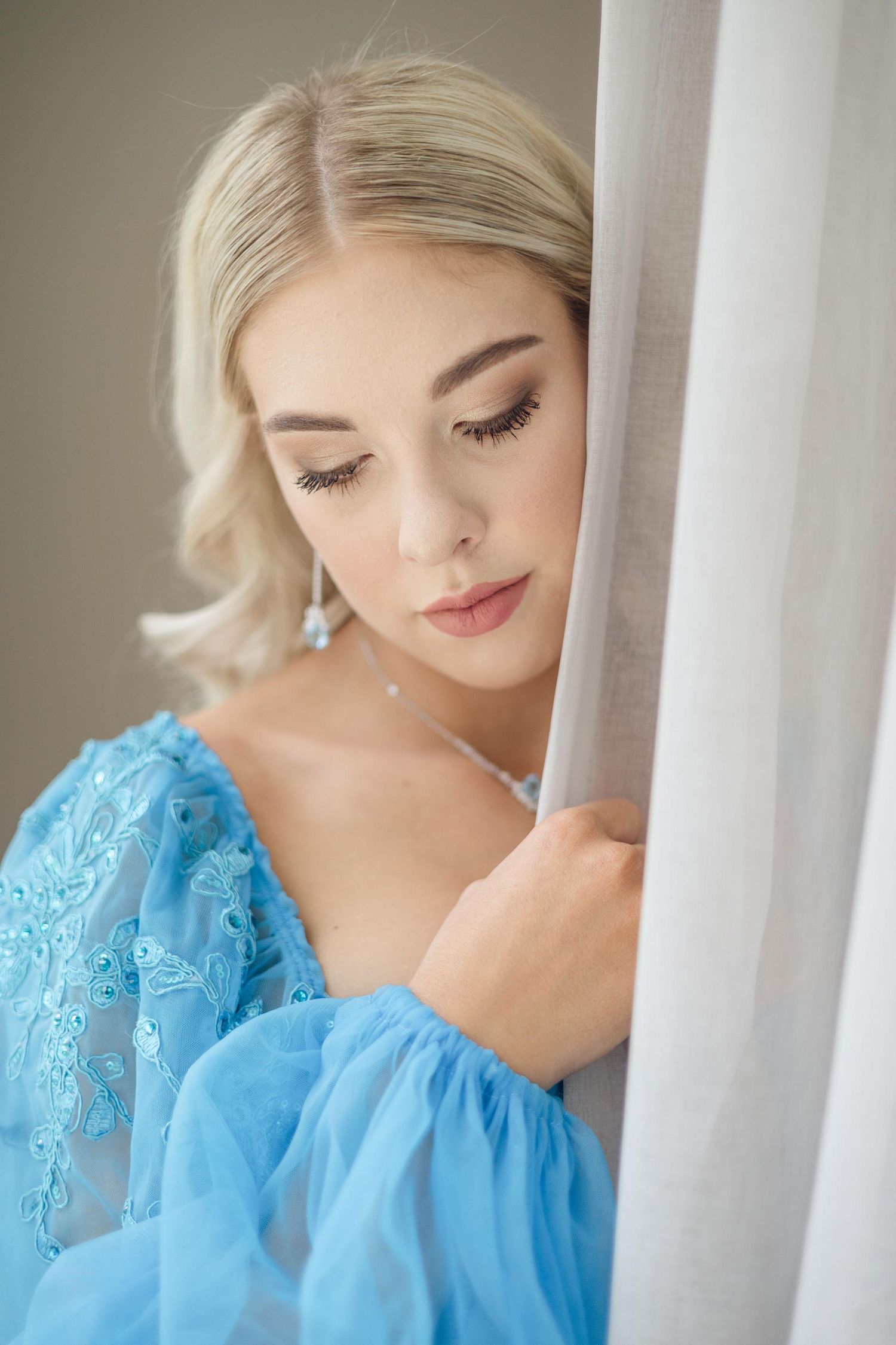 Blonde woman in a light blue lace dress with puffed sleeves gazes downward while leaning against a sheer white curtain.