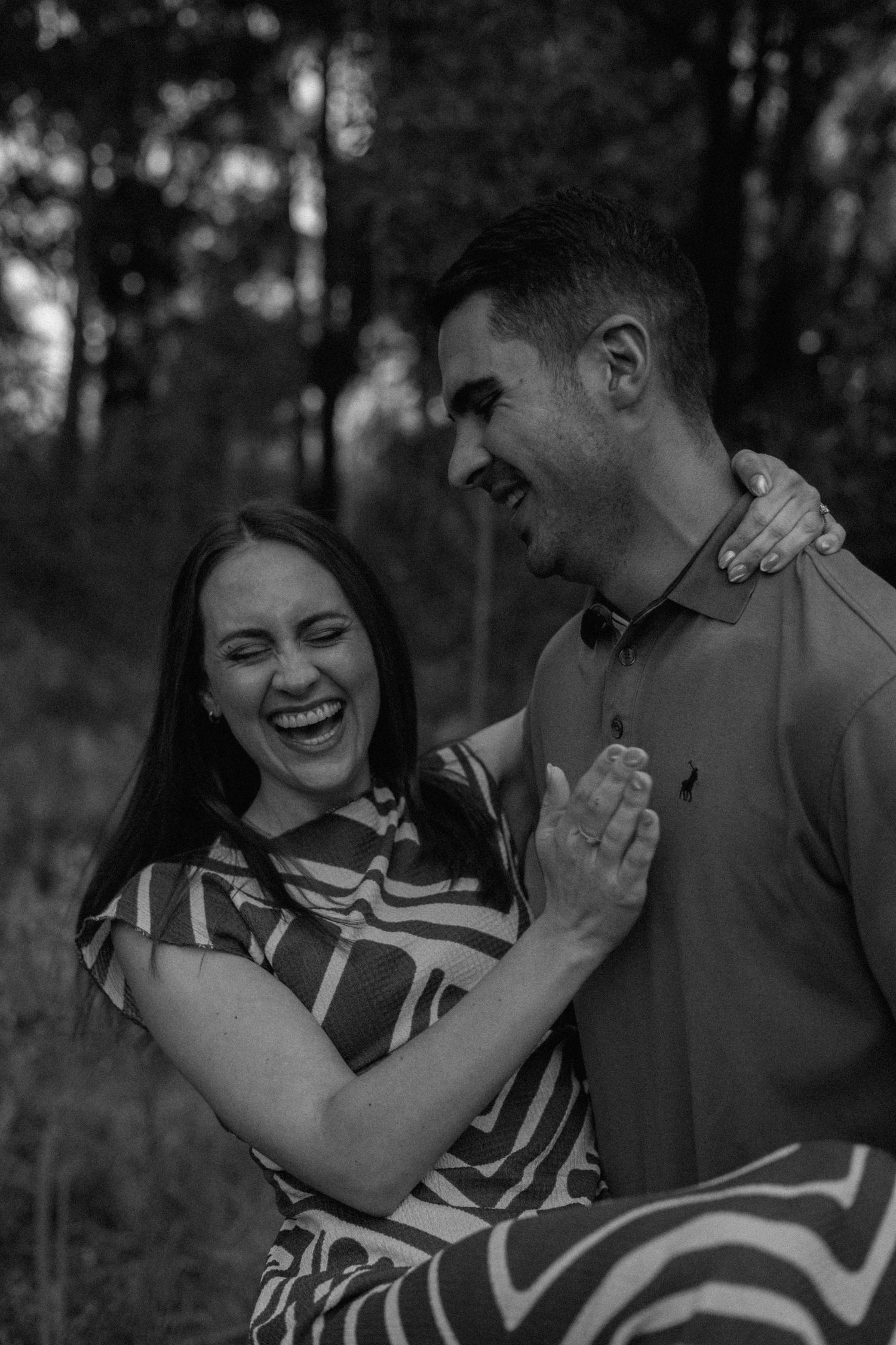 Black and white photo of a joyful couple laughing together in a woodland setting, the woman in a striped dress.