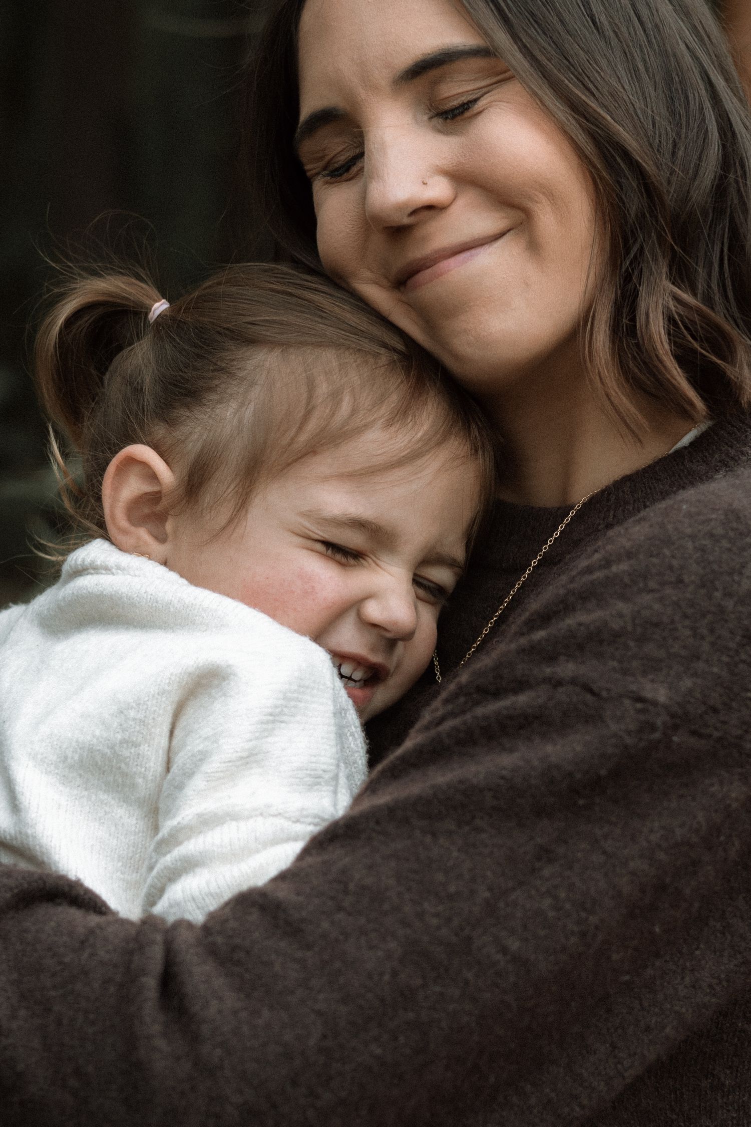 Smiling mother in a brown sweater hugs her giggling toddler daughter in a white jacket during an outdoor lifestyle photo shoot.