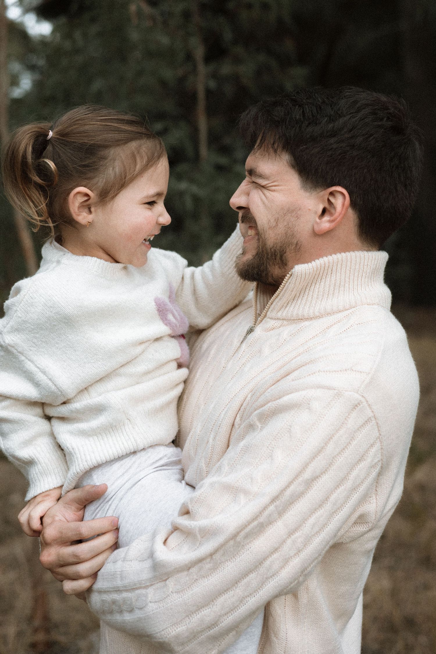 Smiling bearded father in a cream knit sweater holds his laughing young daughter in an outdoor family portrait.