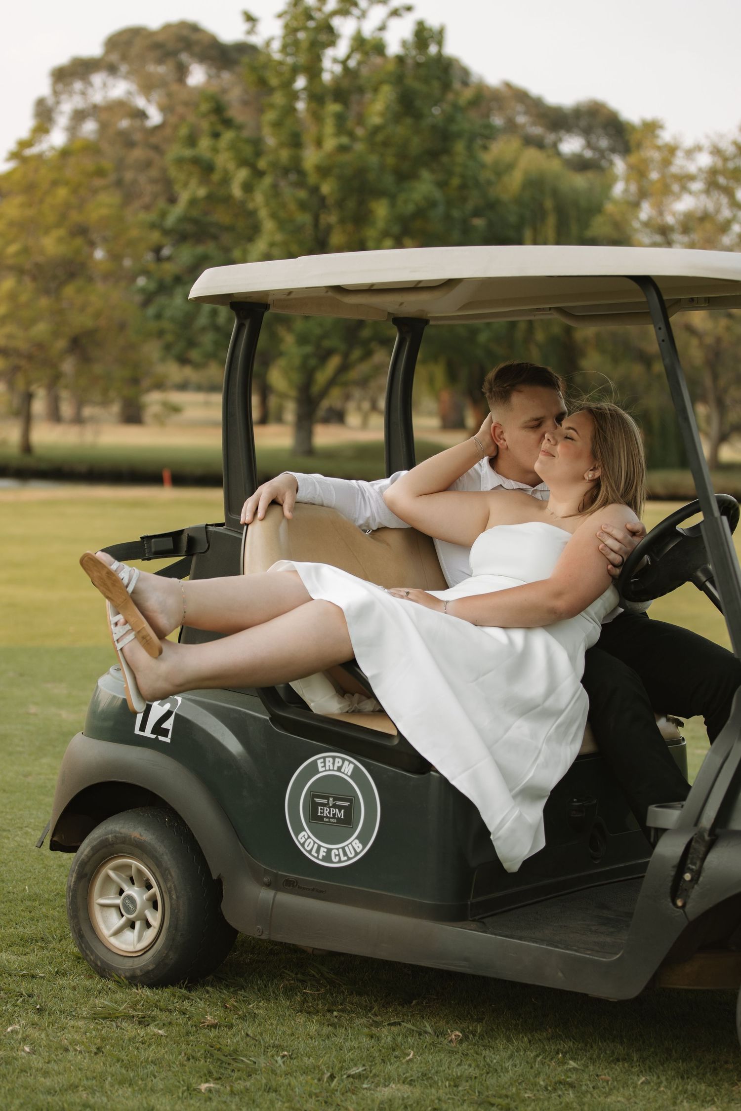 Couple embracing and kissing on a golf cart at a golf club, the woman in a white dress, in a romantic outdoor photoshoot.