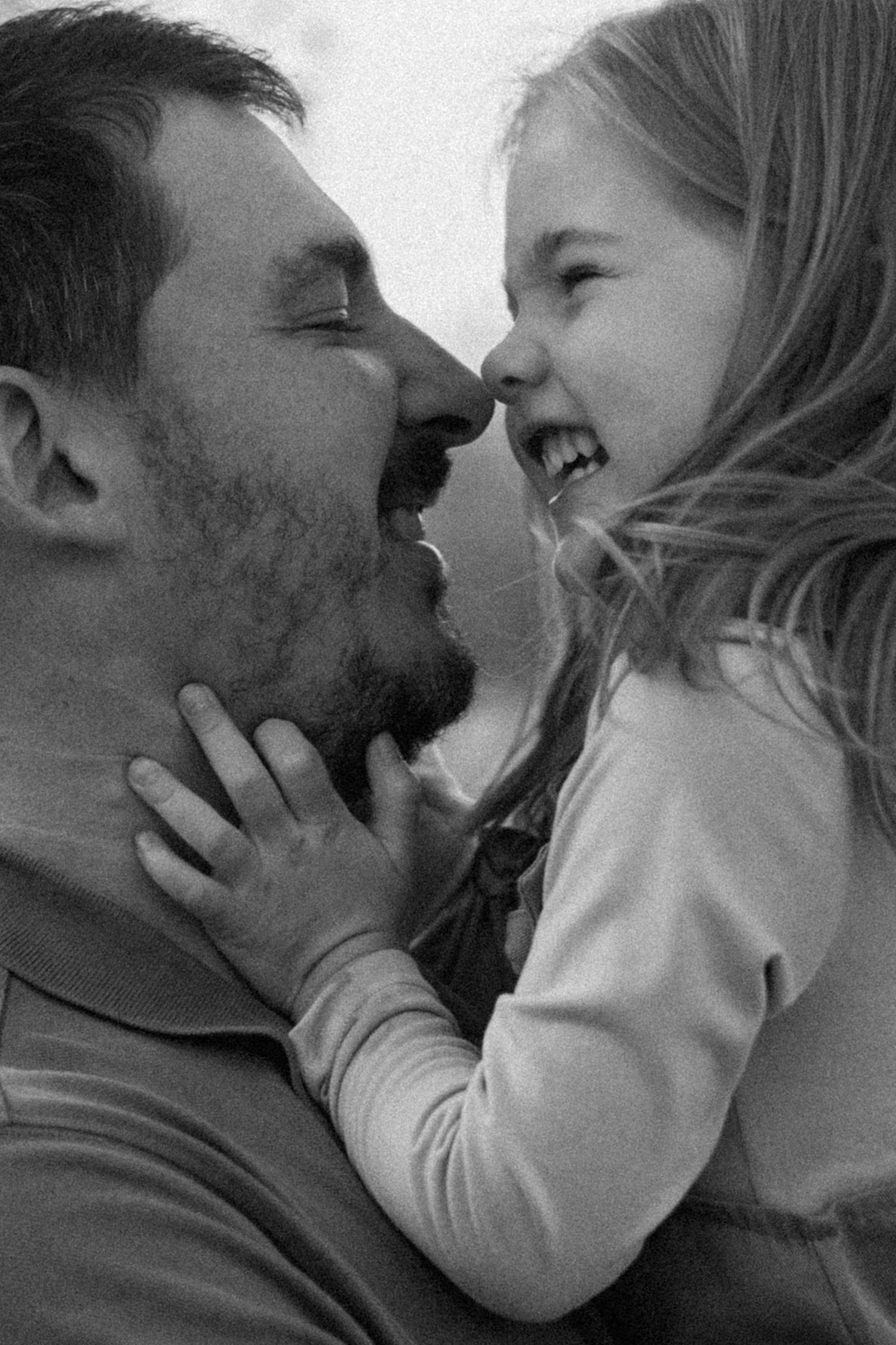 Black and white close-up of a bearded father and his laughing young daughter touching noses in a joyful moment.