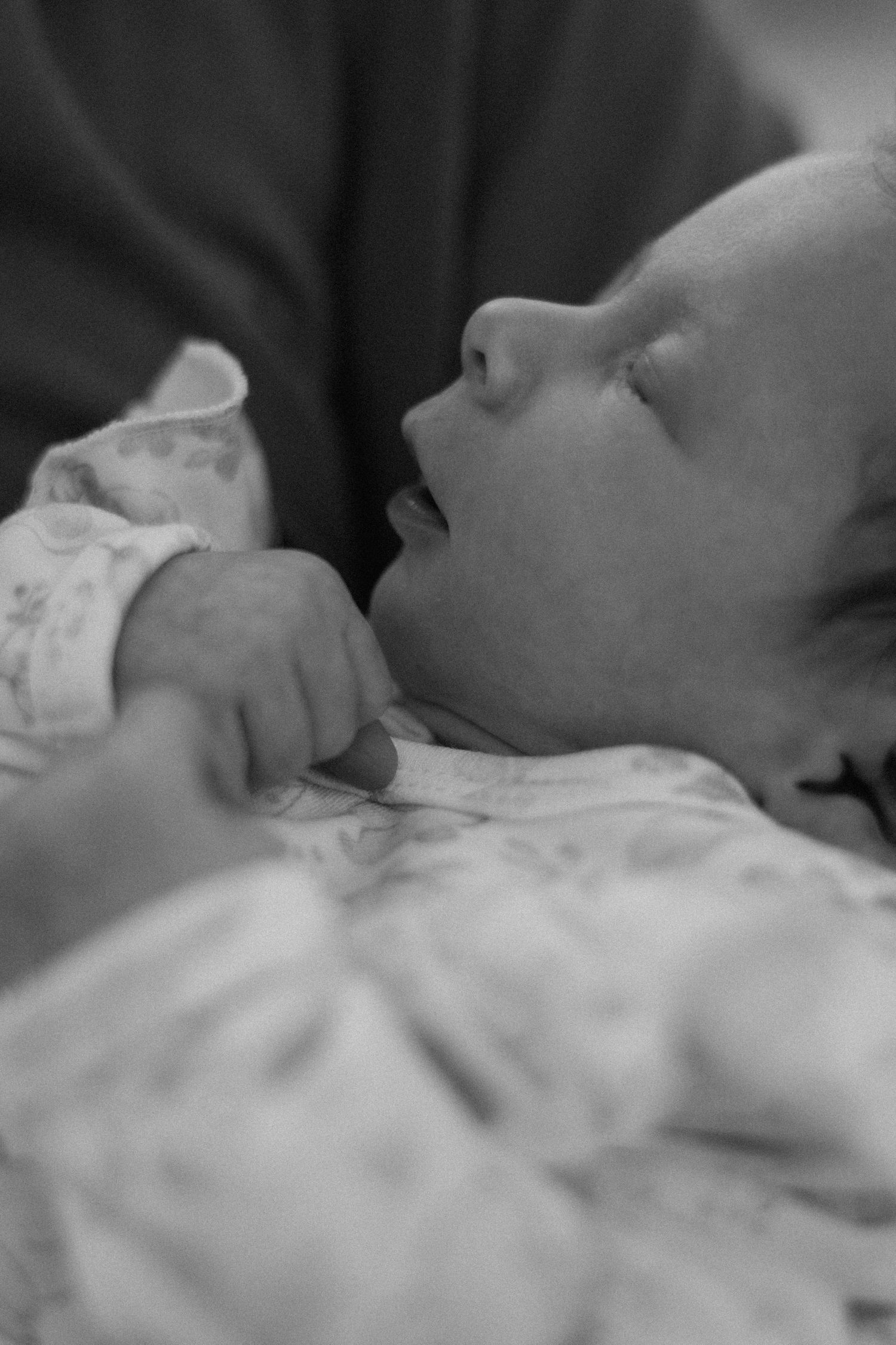 Black and white close-up of a peaceful newborn baby in a patterned onesie with a tiny hand resting near its chest.