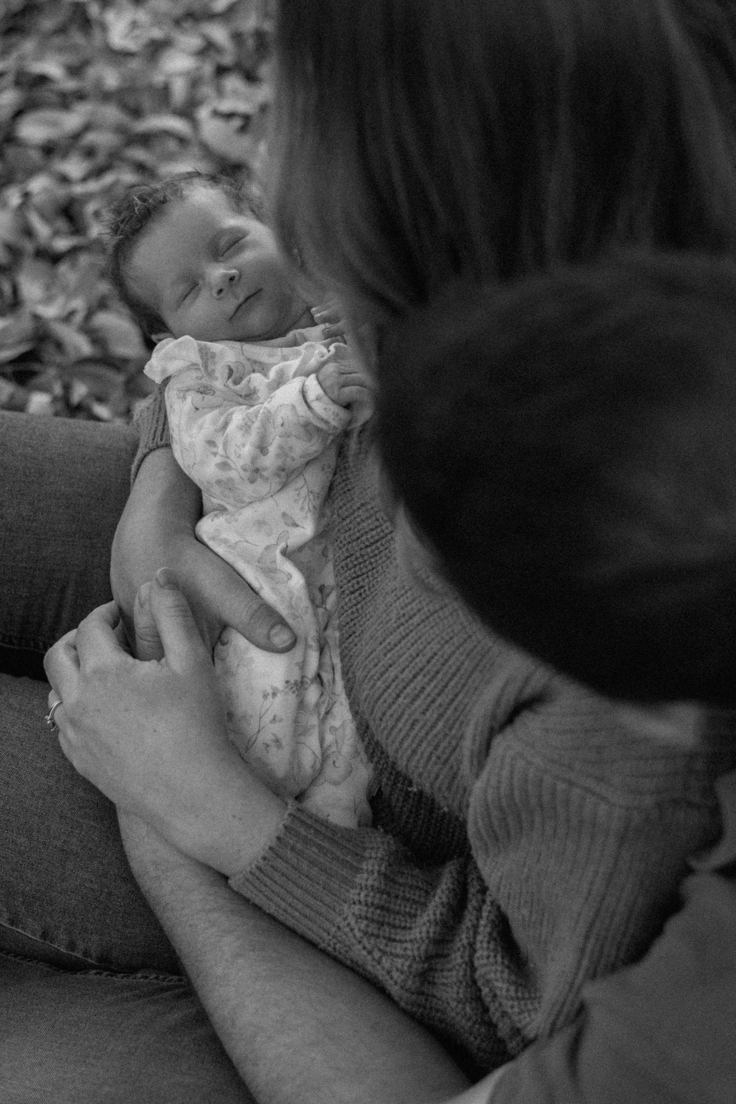 Black and white photo of a sleeping newborn baby wrapped in a soft floral outfit, cradled in a mother's arms outdoors.