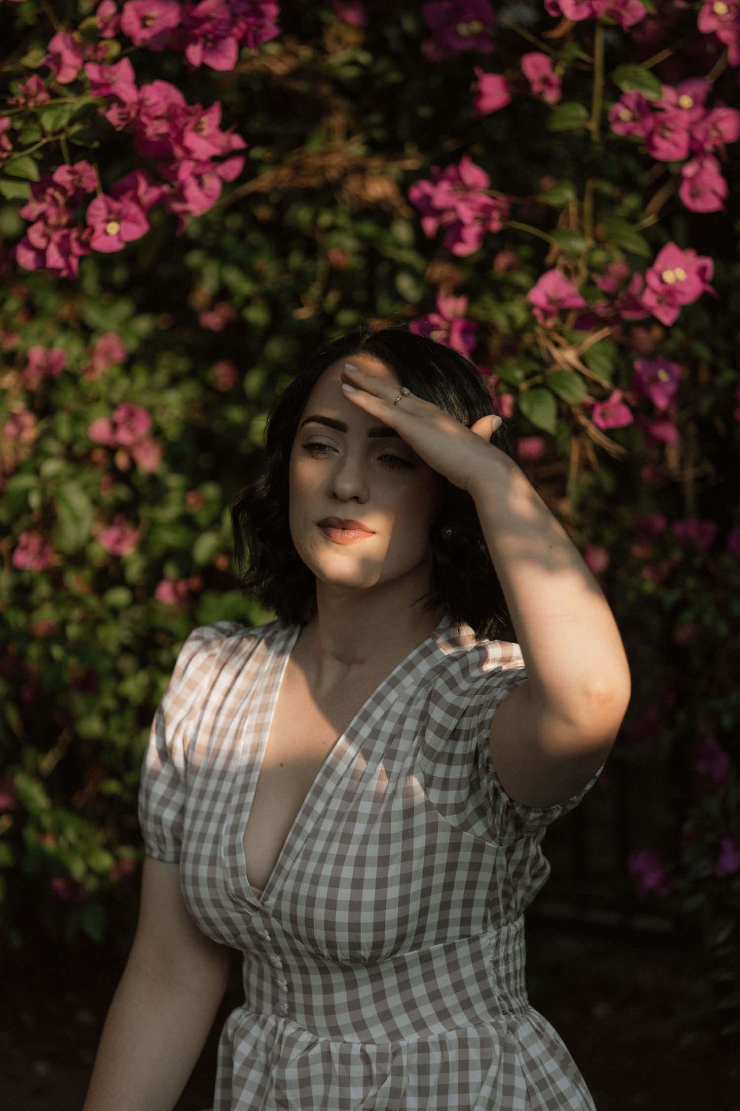 Woman in a gray gingham dress shields her eyes from the sun with her hand in front of blooming pink bougainvillea.