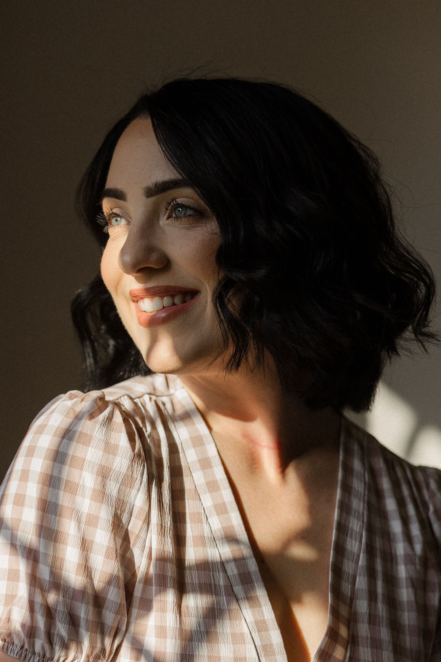 Smiling woman with short wavy black hair in a pink gingham puff-sleeve blouse gazes aside in warm, golden window light.